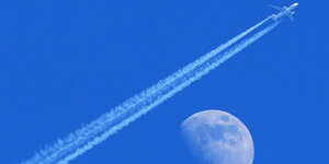 Ein Flugzeug fliegt durch strahlend blauen Himmel und hinterlässt einen Kondensstreifen quer über das gesamte Bild. Im Hintergrund ist der Mond groß zu sehen. Es sieht so aus, als würde das Flugzeug am Mond vorbeifliegen.