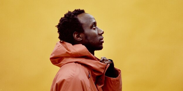 Der Musiker KMRU steht vor einer gelben Wand. Er trägt einen orangenen Regenmantel und ist im Profil zu sehen. Er blickt in Richtung der oberen rechten Bildecke.
