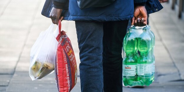Eine Mann trägt seine Einkäufe, in der einen Hand einen roten Beutel und eine Plastiktüte, in der anderen Hand einen Sixpack mit PET-Flaschen