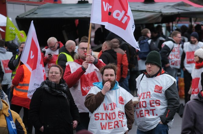 Teilnehmer einer Kundgebung versammeln sich mit Verdi-Fahnen und tragen Überzieher auf denen steht: WIR SIND ES UNS WERT