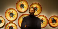 Der Klangkünstler Emeka Ogboh steht in warmen Licht vor einer Wand. An der Wand hinter ihm hängen acht Subwoofer, die in gelbe, bunt bemalte Teller mit roten Rändern eingelassen sind.