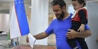 Ein Mann in blauem Shirt hält sein Kind auf dem Arm, gleichzeitig wirft er einen Wahlzettel in eine Wahlurne
