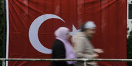 Zwei Frauen laufen durchs Bild, ihre Körper und Gesichter sind unscharf. Hinter ihnen ist eine metergroße Türkeiflagge. Sie ist rot und es ist ein Halbmond sichtbar.