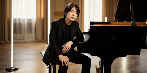 Der Pianist Seong-Jin Cho sitzt an einem Flügel. Er trägt einen schwarzen Anzug und ist zur Kamera gedreht. Sein linker Arm ruht auf dem Klavier neben den Tasten. Das Zimmer, in dem er sitzt hat einen Dielenboden, die Fenster werden von durchscheinenden sowie blickdichten beigen Vorhängen gerahmt.