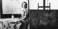 Anna-Eva Bergmann steht in ihrem Atelier an einem Tisch, hinter hier ein von ihr gemaltes Bild