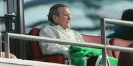 Gerhard Schröder sitzt mitz einer grünen Decke auf einem roten Stadionsitz