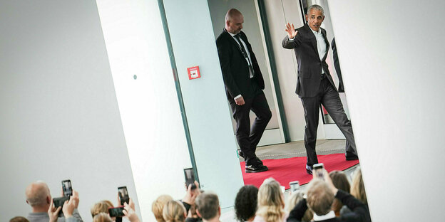 Barack Obama, gefolgt von einem Sicherheitsmann, winkt einer Gruppe von Menschen, die ihn mit dem Smartphone fotografieren