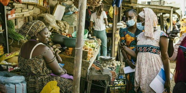 Zwei Frauen mit Gesichtsmaske sprechen auf einem Markt mit einer Marktfrau, die an ihrem Gemüsestand sitzt