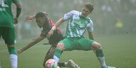 Ein Fußballspieler im grün-weißen Trikot drängt einen Gegner im schwarz-roten Trikot ab