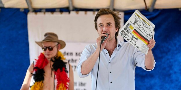 Hendrik Sodenkamp auf einer Bühne mit Mikro und einer Ausgabe des "Demokratischen Widerstands"