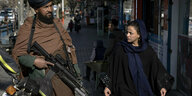 Ein Frau mit Kopftuch und ohne Burka blickt misstrauisch einen schwerbewaffneten Taliban-Kämpfer an.