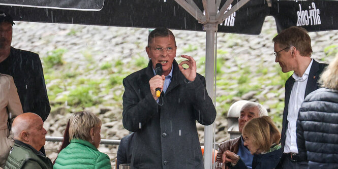 Ein Mann steht bei einer Wahlkampfveranstaltung unter einem Schirm und spricht in ein Mikrofon, es regnet