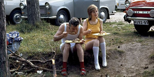 Ein mann und eine junge Frau sitzen in Unterwäsche auf einem Rasen neben einem Parkplatz der DDR