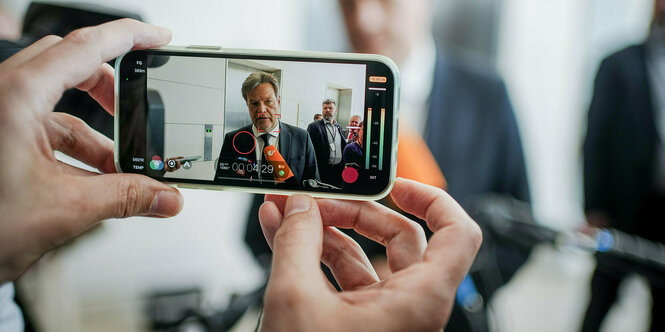 Robert Habeck spricht in die Mikrofone von Journalisten, gesehen durch das Display eines Smartphones