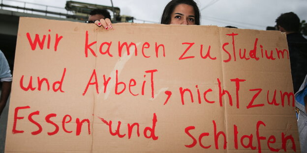 Eine Flüchtlingsfrau in einer Hamburger Flüchtlingsunterkunft hält ein Schild hoch „Wir kamen zu Studium und Arbeit, nicht zum Essen und Schlafen“