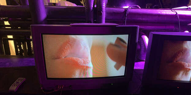Vor einer lila beleuchteten Bühnenkulisse steht ein Fernseher, auf dem ein geschminkter Frauenmund zu sehen ist, der Zigarettenrauch ausbläßt.