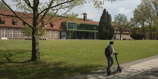 Ein Mann mit eine Rucksack fährt auf einem Roller vor einem Gebäude vorbei