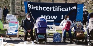 Schwarze Mütter sitzen auf Stühlen auf der Straße und halten ein Transparent hoch, auf dem steht: Birthcertificates now!