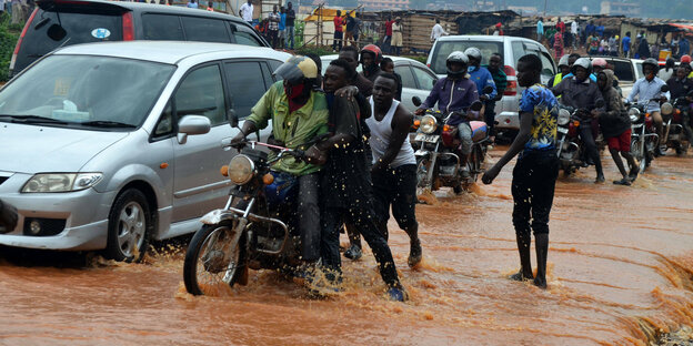 Verkehrszene in der mehrere Männer versuchen ein Mottorad anzuschieben. Die Strasse ist überflutet.