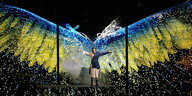 Aljoscha auf einer Bühne vor der Projektion blau-gelber Flügel