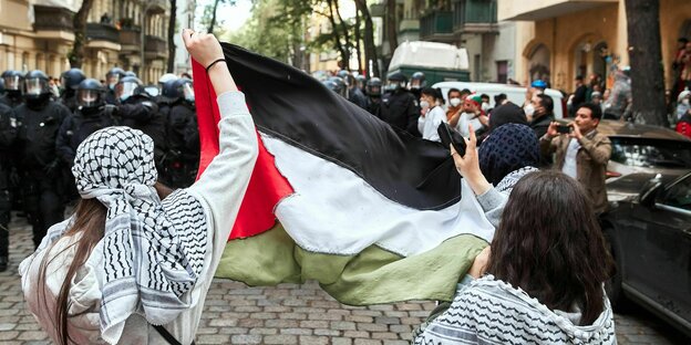 Zwei Frauen halten eine Palästinener-Flagge und stehen einer Reihe von Polizist*innen gegenüber