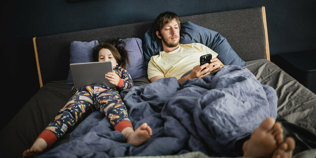 Ein Mann liegt mit smartphone im Bett, daneber ein Kind mit Tablet und buntem Schlafanzug