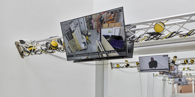 Ausstellungsansicht: An Querbalken sind Bildschirme befestigt, die Überwachungskameraaufnahmen zeigen