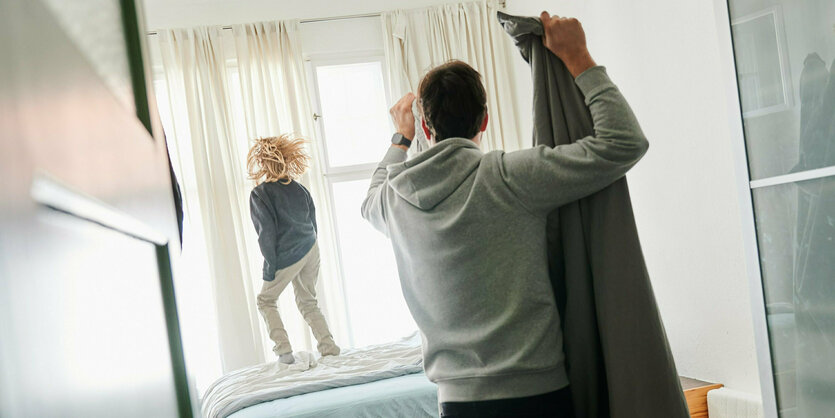 Ein Kind hopst auf dem Bett, ein Mann steht mit Bettwäsche im Raum