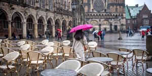 Eine Frau läuft unter einem Regenschirm zwischen den leeren Sitzplätzen eines Cafés auf dem Bremer Marktplatz entlang.