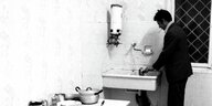 Ein Mann steht im Anzug und Zigarette im Mund an einem Waschbecken in der Küche und wäscht ab