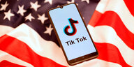 Eine gewellte US-Flagge mit dunklem viereckt voller weißer Sterne und drumrum rot-weiße Streifen. Darauf liegt ein Handy, das das Logo von tiktok sowie den Schriftzug "tik Tok" zeigt.