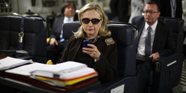 Hillary Clinton sitzt mit Sonnenbrille in einem Flugzeug und schaut auf ihr Handy