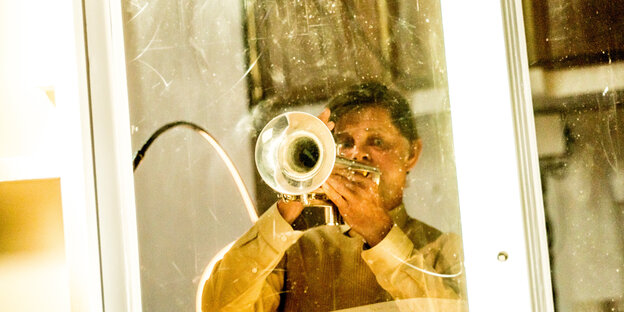 Der Schauspieler Lars Rudolph steht hinter einer Fensterscheibe und spielt Trompete.