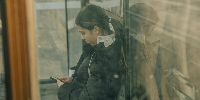 Hinter einer Glaswand hat eine Jugendliche ein Smartphone in der Hand