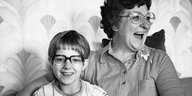 Eine Frau und ein Kind sitzen auf einem Sofa. Die Frau trägt eine Brille und lacht, sie schaut nach rechts. Sie hat den Arm um das Kind gelegt, das ebenfalls eine Brille trägt und in Richtung Kamera lacht. Die Tapete an der Wand ist mit abstrakten fächerförmigem Blattwerk gemustert.