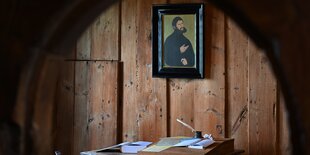 Auf dem Bild ist ein Arbeitszimmer mit hölzerner Wand zu sehen. Unten im Bild ein Schreibtisch mit Papier und Feder. An der Wand ein Portrait des Reformators Martin Luther