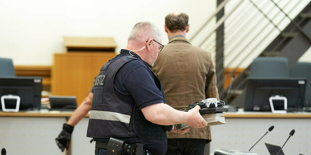Ein Polizist trägt Akten in der hand vor ihm geht ein Mann, man sieht ihn von hinten