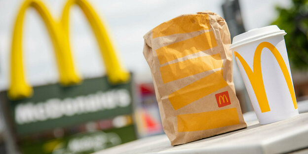 McDonald's To go-Tüte und ein dazugehöriger To go-Becher stehen vor einer McDonald's-Filiale.