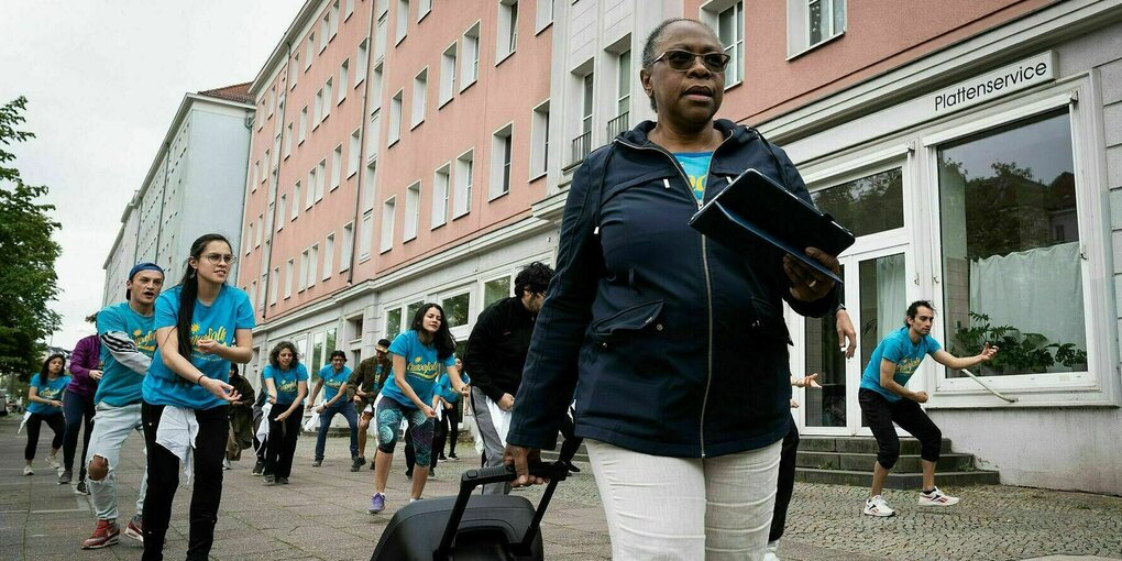 Die Mitglieder der Gruppe Canoafolk üben ihre Performance in blauen T-Shirts aufdem Gehsteig, die Trainerin geht mit der Musikbox vorne weg