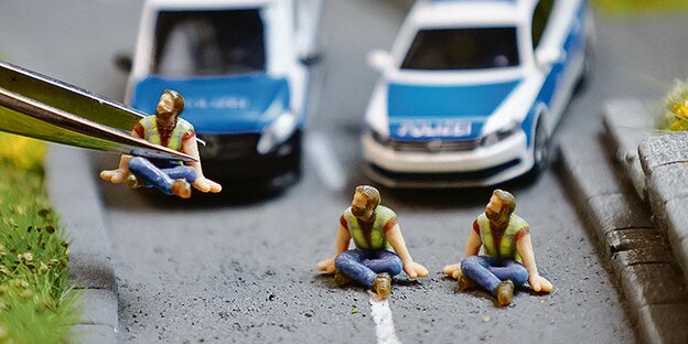 Knetfigürchen Letzte Generation auf einer Straße, dahinter zwei Polizeiwagen