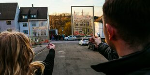 Zwei Personen halten das Bild eines ausgebrannten Hauses vor eine Baulücke