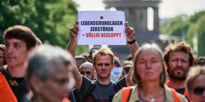 Eine Protestmenge läuft zusammen, sie tragen orangene Warnwesten. Ein Mann in der Mitte hält ein Schild hoch. Drauf steht: "Lebensgrundlage zerstören = völlig bekloppt"