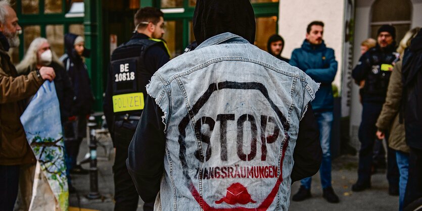 Ein Mensch ist von hinten zu sehen. Auf seiner Jeans-Weste steht "STOP ZWANGSRÄUMUNGEN". Im Hintergrund stehen Polizisten und andere Demonstranten.