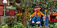 Die Sesamstraßen-Puppe Elin sitzt in ihrem Rollstuhl in einer Sesamstraßen-Kulisse.