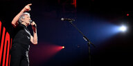 Roger Waters auf der Bühne. Er singt in ein Mikro und zeigt mit einem Finger in Richtung Publikum