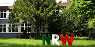 Ein einstöckiges Gebäude mit grauer Fassead hinter Bäumen. Davor steht eine Skulptur bestehend aus den Buchstaben N, R und W - gehalten in den Farben des Wappens von NRW