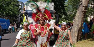 Zwei Damen stehen links und rechts um einen Mann und lächeln in die Kamera. Der mann trägt ein Rot-Goldenes Karnevals-Kostüm mit Federn und Kopfschmuck. Die beiden Frauen traditionelle weiße Kleider aus Brasilien mit bunten Schürzen.