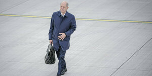 Olaf Scholz in dunklem Anzug. Er geht alleine über das Rollfeld des Berliner Flughafens BER. Er trägt eine Handtasche.