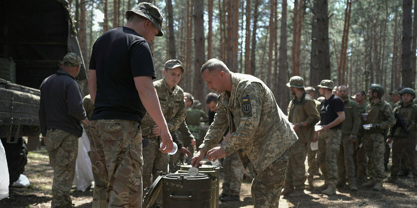 Soldaten im Wald stehen Schlange, um sich mit einer Tasse Suppe aus einem großen Kessel zu schöpfen