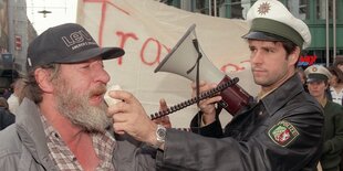 Der Regisseur und Künstler Christoph Schlingensief interviewt als Polizist verkleidet in der Hamburger Innenstadt einen Obdachlosen.